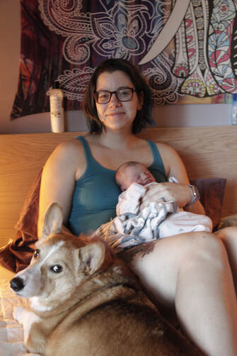 Newly postpartum person cuddling in bed with new baby and corgi.